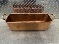 Side view showing rectangular brass container with two handles on opposite shorter sides, placed on a concrete floor with a diamond plate wall in the background.