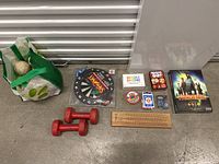 Photo showing multiple game-related items including Pandemic board game, Jungle Speed card game, Cribbage board, dartboard, red dumbbells, and a green bag.