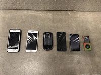 Wide shot of six Apple devices including iPhones and iPods arranged in a horizontal line on a metallic surface, with most screens cracked or damaged.