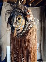 Front and slightly angled view showing the head covering mask with painted wooden face, cowrie shells, hair strands on top, and long fiber fringe