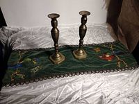 Pair of heavy antique brass candlesticks standing on green embroidered textile runner with birds and butterflies, bordered with decorative trim.