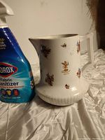 Full side view of the white porcelain pitcher with floral designs and Brown Palace Denver logo, next to a fabric sanitizer bottle to show scale.