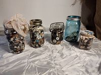 Front view of five jars filled with assorted vintage buttons in various sizes and colors on a white cloth