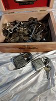Wooden box filled with many antique keys and key parts along with two metal padlocks placed on white fabric.