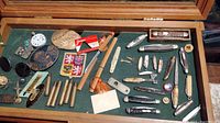 Full view of mixed vintage items including pocket knives, patches, collectibles, and other small vintage pieces laid on green surface inside a display case.