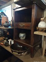 Front and side view of the vintage wooden cabinet showing single brass-handle drawer and open shelving compartments, surrounded by pottery items not included.