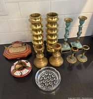 Lot of brass candle holders, Moroccan style pen holder, and two ashtrays shown on black countertop