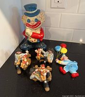 Photo of a large vintage clown coin bank with blue hat and several small clown figurines around it.
