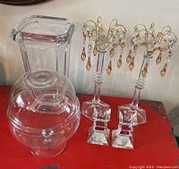 Photo of 2 tall crystal candle holders with hanging amber crystal teardrops, one tall rectangular glass vase, and round glass candy jar with lid on red surface