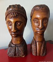Two hand carved wooden African bust sculptures shown side by side, displaying carved facial details and clothing patterns.