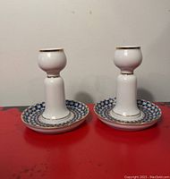 Two white porcelain candle stick holders with blue and gold detailed base pattern and gold trim, placed on red surface