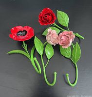 Four painted metal flower wall hooks; a red poppy, red rose, and two pink roses, each with attached green metal leaves and hooks. All laid flat on black background.