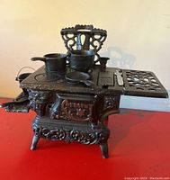 Front side of miniature cast iron stove showing ornate details, burners, and utensils on top and sides.