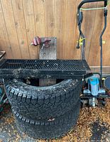 Two large off-road tires stacked with black metal folding ramp resting on top next to a blue electric garden tiller tool against wood wall