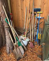Various gardening tools leaning against a wooden fence and tree. Includes shovels, rakes, broom, cultivator, garden fork, hoe, bucket with ropes.