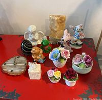 Photo of various mostly vintage decorative items including Murano glass egg paperweight, porcelain china flowers, carved stone animal figurine, small clock, and figurines arranged on red surface.