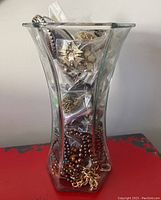 Full view of the 13in tall clear glass vase filled with assorted costume jewelry in plastic bags and loose chains and beads at the bottom.