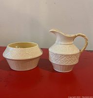Set of two porcelain items on a red surface; cream pitcher on right with gold-accent detail and sugar bowl on left.