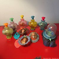 Collection of eight vintage Little Kiddle dolls in various colorful perfume bottle-shaped containers and decorative frames, displayed on a red surface.