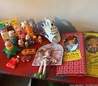 Multiple vintage toys including spaceship, dolls in boxes, and various plastic figures arranged on red table.