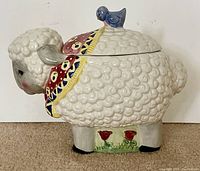 Side view of ceramic lamb and blue bird cookie jar showing intricate wool texture, colorful collar, blue bird on lid, and floral details near legs.