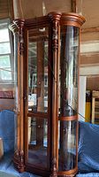 Lighted solid wood curio cabinet with carved front columns, curved glass panels, and multiple glass shelves inside.