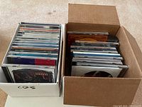 Two boxes filled with 30 assorted music CDs in jewel cases showing edges of covers and backs