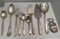Overview of all 10 pieces showing a variety of spoons, forks, serving spoon, cake server and metal bottle opener. Pieces are tarnished and need cleaning.