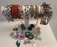 Bracelets of various styles on white jewelry stand with multiple pairs of earrings and necklaces arranged below.