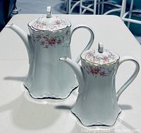 Front view of two porcelain teapots showing floral pattern and gold trim, placed on a table.