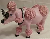 Side view of the Cow Parade French Moodle resin figurine showing pink body, textured poodle-like fur areas, black nose, blue eye, studded collar, and bow.