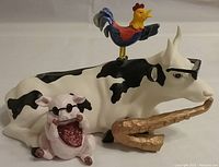 Resin figurine of cow laying down, rooster on cow's back, pig with sunglasses and bandana beside cow, holding a gold saxophone-shaped object
