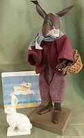 Full view of the Shadow Dancer rabbit with cloth clothes, metal legs, wood base, white Dept. 56 Snowbunny figurine, and ceramic trivet with rabbit painting.