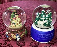 Front view of two Christmas snow globes on red patterned tablecloth showing snowman globe with decorative painted base and Nelvana polar bear globe with blue base and stars