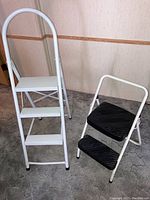 Two foldable step ladders side by side against wall and carpet; one black and white, one white aluminum