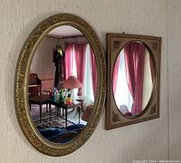 Two hallway mirrors mounted on wall with reflections showing room interiors. The left mirror is oval with an ornate gold frame; the right is square with an oval mirror and decorative corner accents.