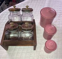 Photo showing four glass canisters with worn wooden lids arranged on a wooden stand and three pink glazed ceramic canisters with lids beside them on a white quilted surface.