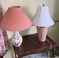 Two table lamps on a small wooden table: one ceramic with cherry blossom design and pink shade, one pink base with white bell shade with brass detail.