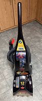Black Bissell upright carpet cleaner front view with sticker indicating '6 cleaning rows', 'new improved', and '12 amps'.