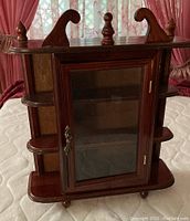Front view of the cabinet showing wood finish, glass door, decorative finials on top, side shelves, and feet.