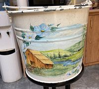 Front side close-up showing the hand-painted country scene with barn, river, flowers, and hills on the white metal bucket.