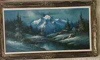 Front view of framed winter landscape painting showing mountain, stream, trees, and cabin in a snow-covered setting.