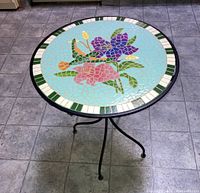 Side view of round side table showing the colorful hummingbird and floral ceramic mosaic top and black metal frame with tripod legs.
