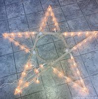 Photo of the star fully unfolded flat on floor with warm white icicle lights attached along edges and connected to an electrical cord with plug extending from the frame.