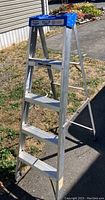 Full side view of the Lite aluminum step ladder standing upright on pavement and next to grass and a building.