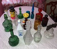 Front view of 16 assorted glass bottles on a table with a white cloth, showing varying colors and shapes including green, clear, blue, yellow, and amber bottles mostly with stoppers.
