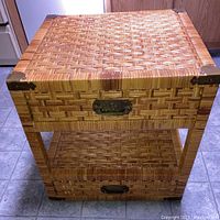 Front view of rattan bedside table showing woven pattern, metal corner reinforcements, metal handles on two drawers, and bottom open tier shelf.