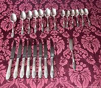 Full layout of the silverware collection on a patterned cloth background, showing spoons arranged in rows and knives below them.