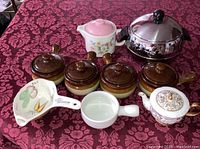 Overview of the ceramic soup bowls, tea pots, gravy separator, and metal soup tureen arranged on a patterned cloth.