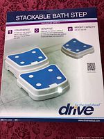 Box front showing stackable bath step with blue non-slip surface and white plastic body. Includes convenience, versatility, and weight capacity info.